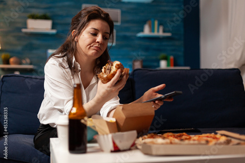 Obraz na plátně Caucasian female holding tastu burgery in hands browsing on social media using smartphone relaxing on couch during junck-food night
