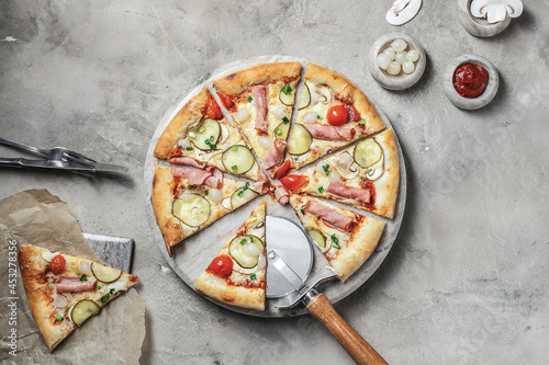 Tasty pizza with ham, cherry tomatoes, mozzarella, mushrooms, green onions and pickle cucumber on marble round tray. Garnished on grey stone backdrop with pizza ingredients