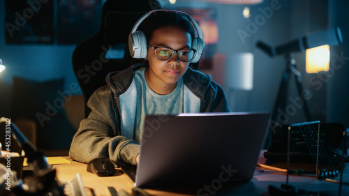 Young Teenage Multiethnic Girl Using Laptop Computer and Wearing Headphones in a Dark Cozy Room at Home. She's Browsing Educational Research Online. Studying Science School Homework Concept.