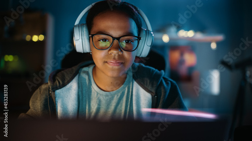 Young Teenage Multiethnic Girl Using Laptop Computer and Wearing Headphones in a Dark Cozy Room at Home. She's Browsing Educational Research Online. Studying Science School Homework Concept. © Gorodenkoff