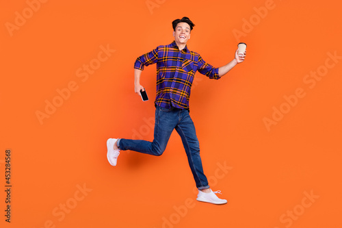 Full body profile photo of young guy jump hold coffee telephone wear shirt jeans sneakers isolated on orange background