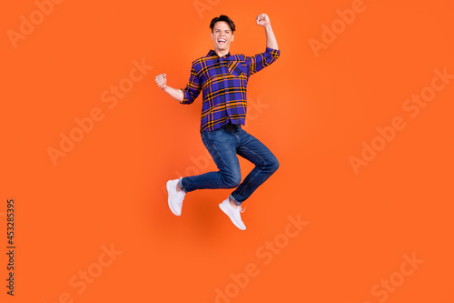 Photo of sweet lucky teenager man dressed checkered clothes jumping high rising fists smiling isolated orange color background
