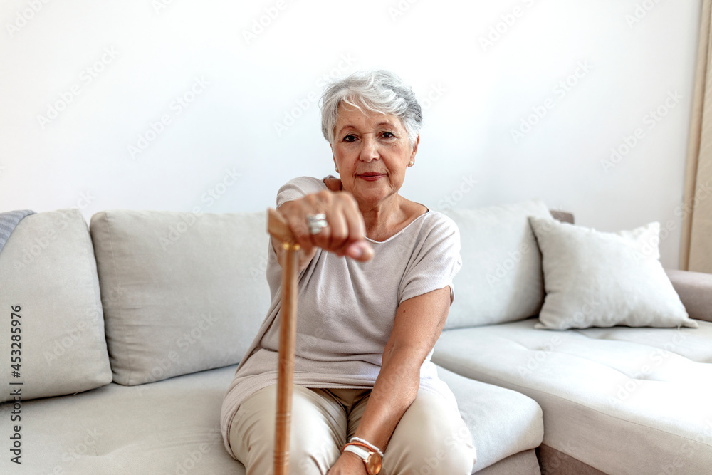 Selective focus of a retired woman with her wooden walking stick at home. Cropped shot of a senior woman using a walking stick at home. Old gray hair Serious lady supporting on a walking cane at home.