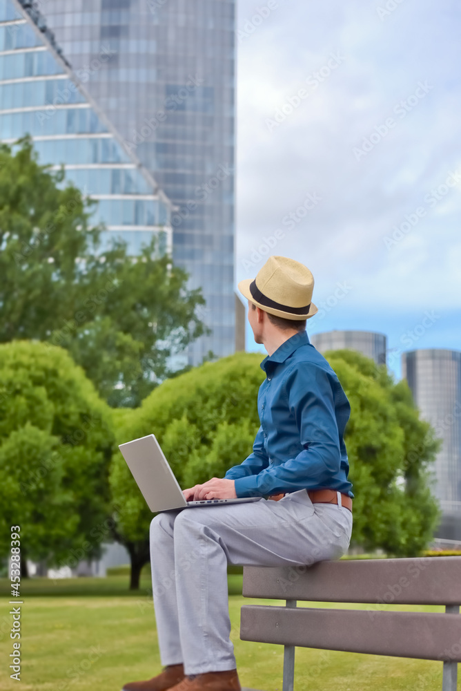 A man with a laptop works remotely on a bench outdoors and looks at an office building.