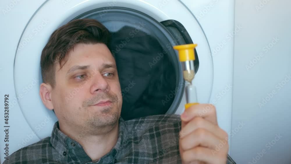 Man with sandglass is waiting the washing machine with grey bedspread inside it, doing his laundry at home or laundry room. Everyday life, housework and household of young man. Looking at sandglass. Man with sandglass is waiting the washing machine with grey bedspread inside it, doing his laundry at home or laundry room. Everyday life, housework and household of young man. Looking at sandglass.