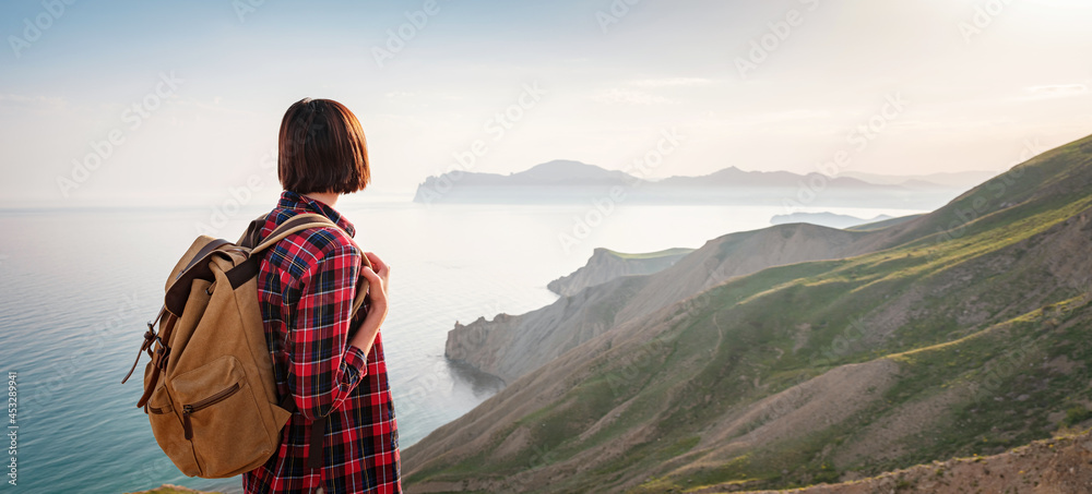 Naklejka premium A young Asian woman with a backpack hiking in the summer enjoying a sunny view of the sea. Mountain and coastal travel, freedom and an active lifestyle