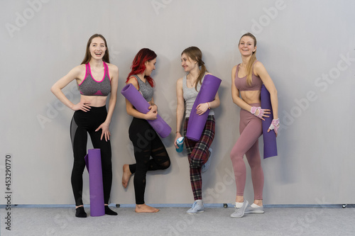 four charming athletic girls in sports out fits talking and smiling standing next to the wall holding a yoga mat, smiling to each other, standing at a grey background. 