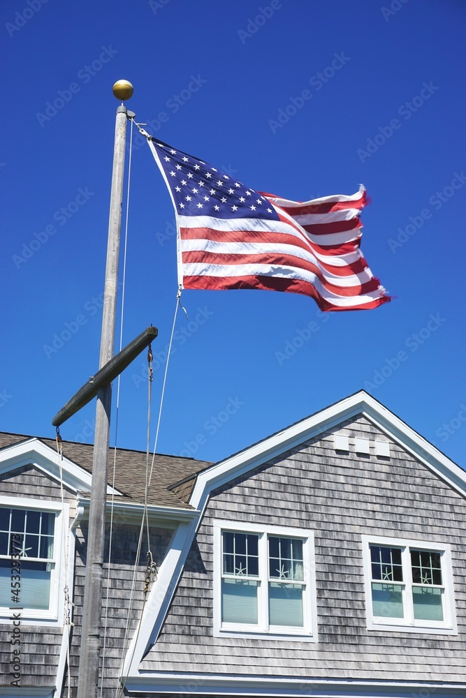 American flag flying on a nautical mast flagpole in front of a typical New England building with gable roof and wooden shake shingles with clear blue sky above