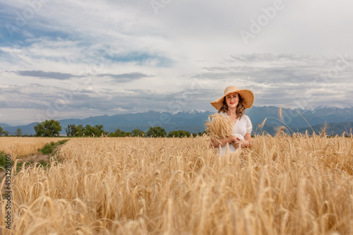 a young attractive woman in a white dress and a straw hat on a field of ripe cereals against a blue sky with clouds in autumn  the concept of harvesting  agribusiness and agriculture