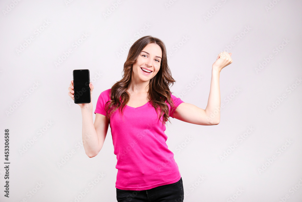 Young beautiful brunette woman with long wavy hair wearing pink shirt, posing with a phone over isolated background. Portrait of female model showing blank screen of a cellphone. Close up, copy space. Young beautiful brunette woman with long wavy hair wearing pink shirt, posing with a phone over isolated background. Portrait of female model showing blank screen of a cellphone. Close up, copy space.