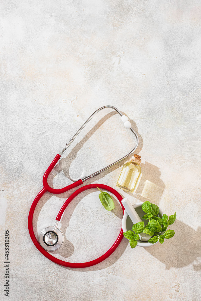 Transparent bottle of basil essential oil, fresh basil leaves in a ceramic mortar with a pestle and stethoscope on a light concrete background. Homeopathy medicine concept.