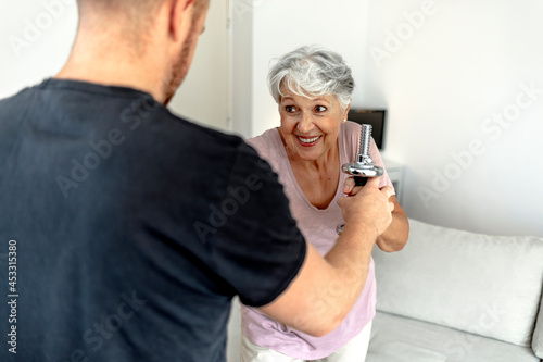 Shot of a fitness instructor assisting a senior woman with some weightlifting exercises. Young male therapist standing next to a senior patient and helping her to lift weights with arm and smiling.