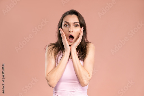 Portrait of shocked young woman wearing pink t-shirt with hands on her face, stun expression on her face with lots emotions. Facial expressions, emotions, feelings.