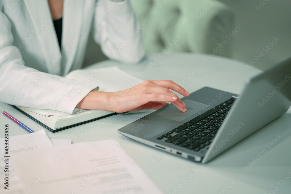 No face visible Businesswoman working sitting in front of laptop wearing white official suit. Office worker female using touchpad browsing laptop while sitting at her working place. Close up. No face visible Businesswoman working sitting in front of laptop wearing white official suit. Office worker female using touchpad browsing laptop while sitting at her working place. Close up.