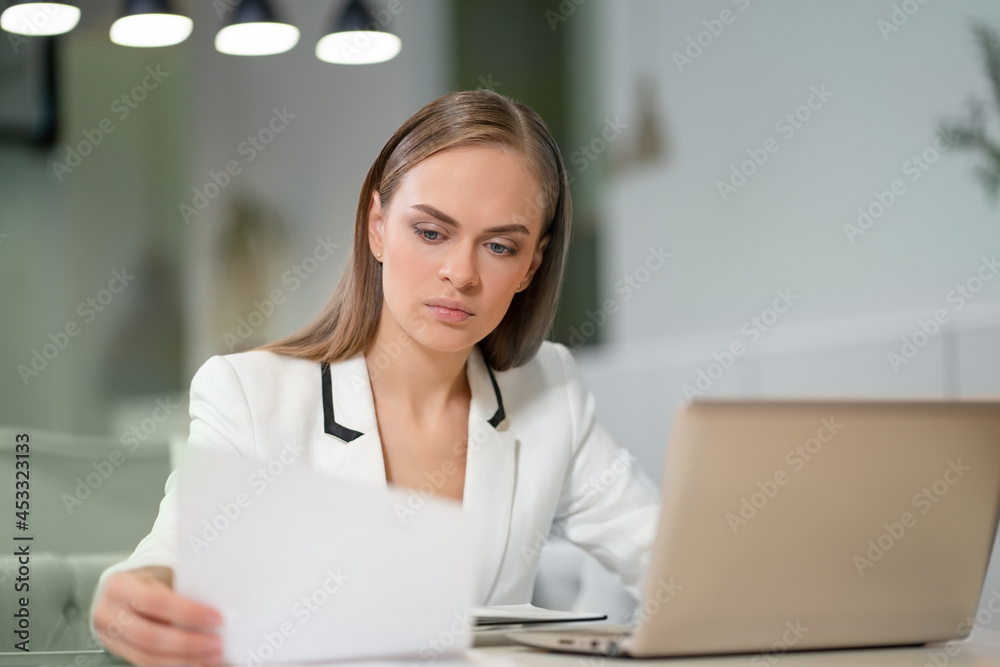 Portrait of a beautiful businesswoman in white jacket working with documents in her hands sitting in front of laptop. Office worker looking at her working place. Portrait of a beautiful businesswoman in white jacket working with documents in her hands sitting in front of laptop. Office worker looking at her working place.