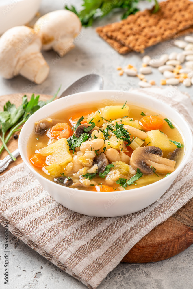 Soup with barley, beans, mushrooms, potatoes, carrots, celery, and herbs. Barley soup with vegetables in a white bowl on a gray concrete background. Vegan food, homemade soup. Soup with barley, beans, mushrooms, potatoes, carrots, celery, and herbs. Barley soup with vegetables in a white bowl on a gray concrete background. Vegan food, homemade soup.