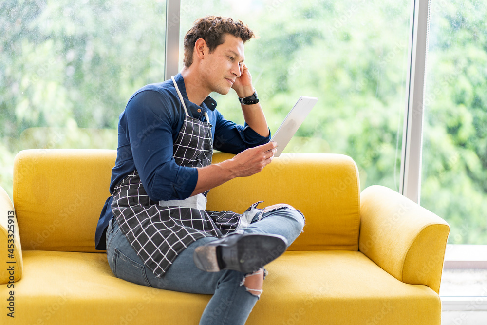 small business owner, wearing apron, sits at work, looks at a tablet, checks online for orders on a yellow sofa in a restaurant. small business owner, wearing apron, sits at work, looks at a tablet, checks online for orders on a yellow sofa in a restaurant.