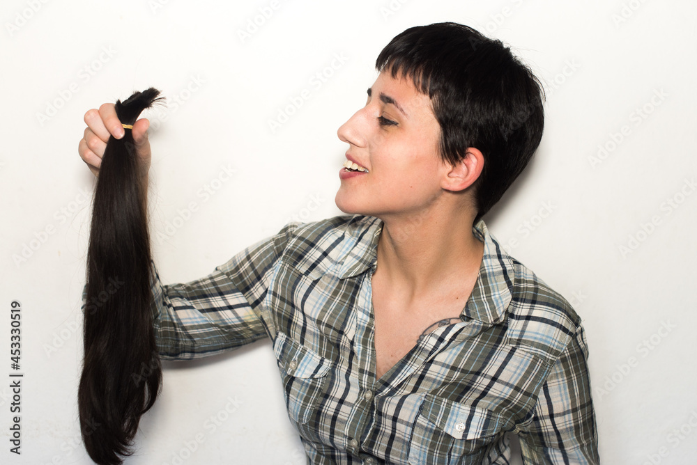 Mujer joven con pelo corto sosteniendo su cabello cortado en una cola de caballo. Concepto de donación de cabello para pacientes con cáncer. - obrazy, fototapety, plakaty 