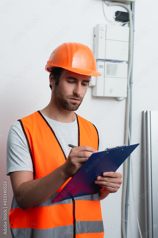 Fototapeta premium Workman in safety vest writing on clipboard near switchboard