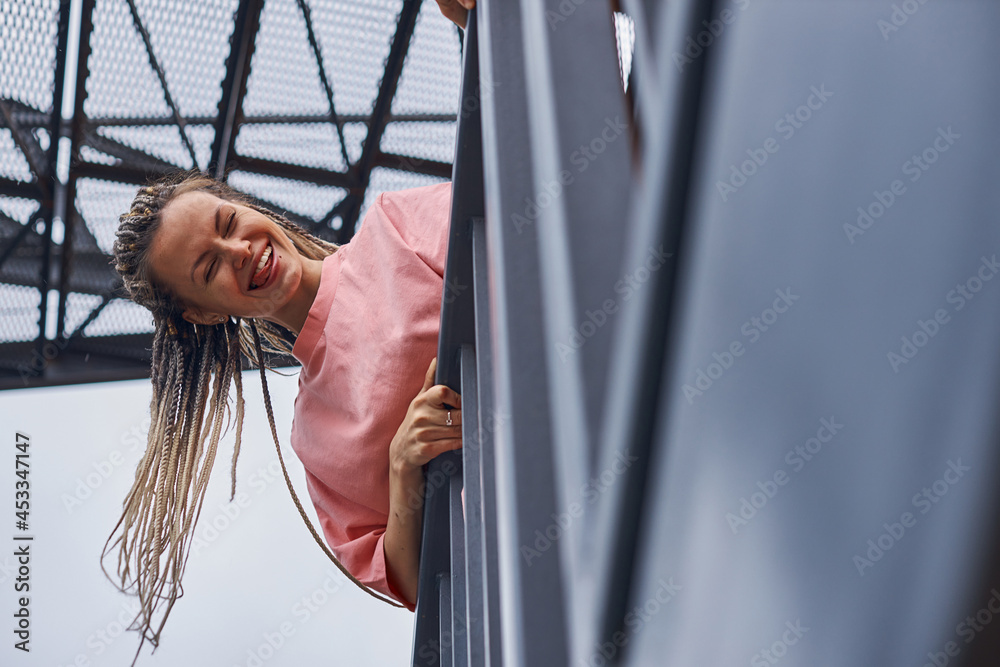 Obraz premium funny young girl laughs and shows her tongue looking down at the camera standing on an iron staircase on the street of the city. High quality photo