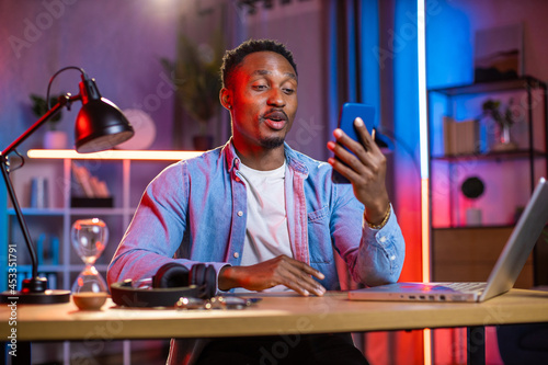 Black male freelancer sitting at home office with laptop on table and talking during video call. Handsome man in denim shirt using modern smartphone for online conversation.