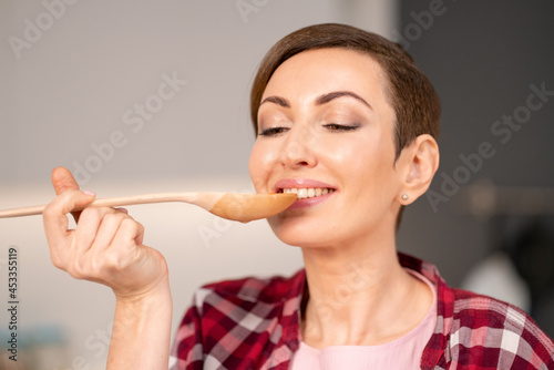 Fototapeta Naklejka Na Ścianę i Meble -  Close-up of a woman trying a cooked dish using a long wooden spoon. Woman with a short hair cooking a dinner for family standing in the modern kitchen of a new house. Healthy food at home. 