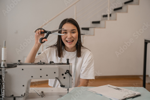 Cheerful asian designer holding scissors near sewing machine  fabric and clipboard