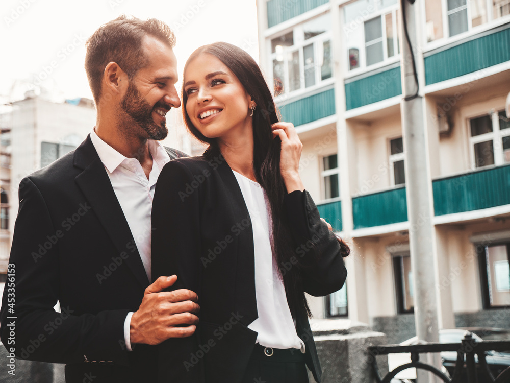 Beautiful fashion woman and her handsome boyfriend in suit. Sexy smiling brunette model in black evening dress. Fashionable couple posing in the street at sunset. Brutal man and his female outdoors Beautiful fashion woman and her handsome boyfriend in suit. Sexy smiling brunette model in black evening dress. Fashionable couple posing in the street at sunset. Brutal man and his female outdoors