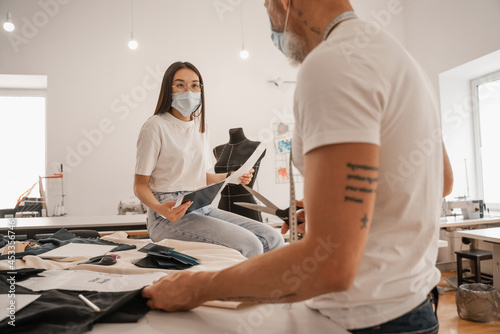 Asian designer holding papers near blurred colleague in medical mask in studio