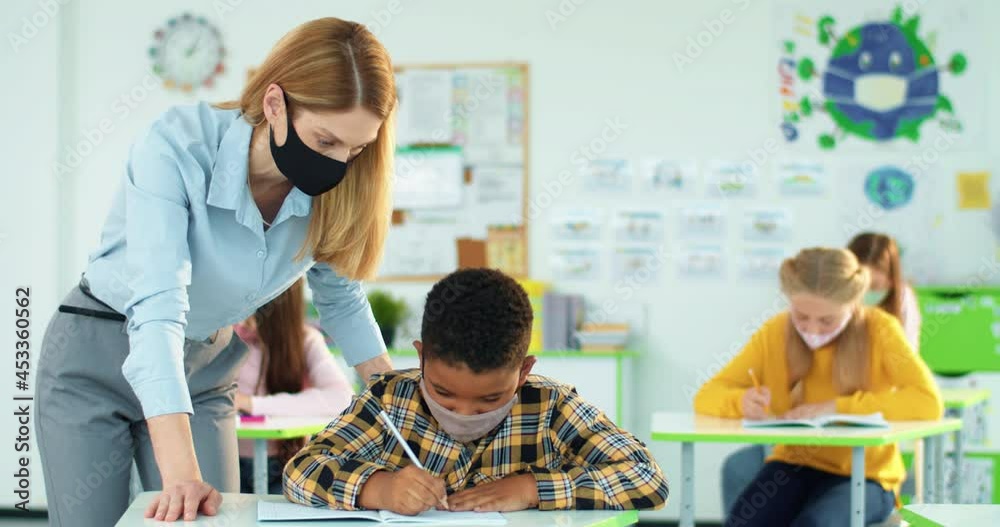 Portrait of young beautiful Caucasian woman teacher helping little African American junior student boy to write test in copybook at class. Female teaching bright schoolboy explaining lesson Post-covid