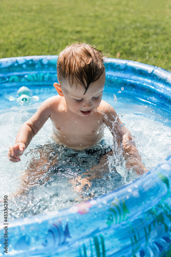 toddler boy sitting in inflatable pool and making water splashes toddler boy sitting in inflatable pool and making water splashes