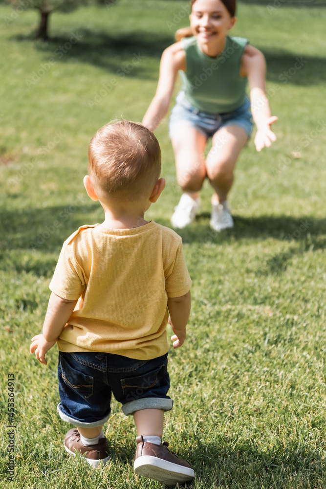 back view of toddler boy near blurred cheerful mother with outstretched hands back view of toddler boy near blurred cheerful mother with outstretched hands