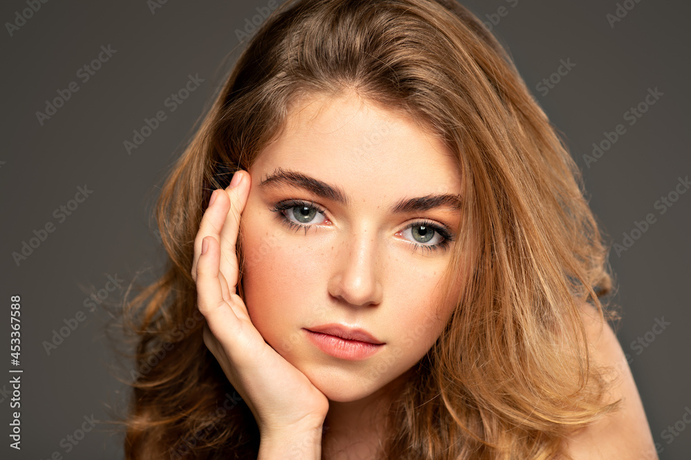 Fototapeta premium Pretty young woman with long brown hair looking at camera. Closeup portrait of an young adult girl with long curly hair. Photo of a fashion model posing at studio. Beauty portrait.