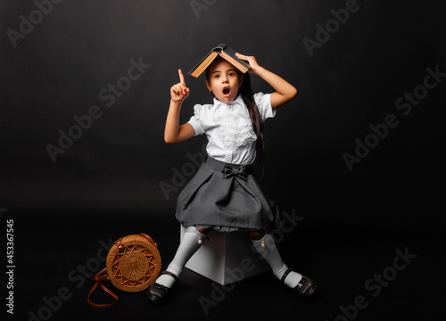 Cute, 7 years old schoolgirl, with a book on her head pointing upwards with an open mouth, is isolated on a dark background. photo