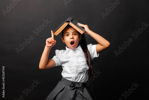 Cute, 7 years old schoolgirl, with a book on her head pointing upwards with an open mouth, is isolated on a dark background. photo