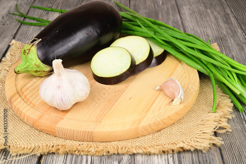A whole eggplant next to another one, chopped into slices, a head of garlic and green onions, laid out on a wooden cutting board