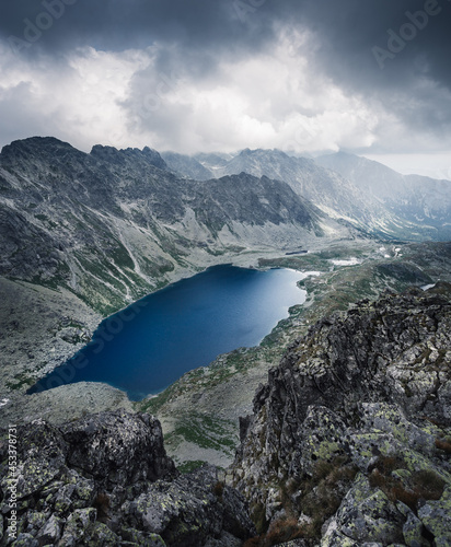 Mountain valley with a lake of glacial origin. The lake is surrounded by rocky mountains and a spring flows from its end towards the valley. The sky is cloudy with dark clouds.