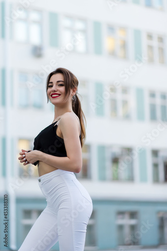 A beautiful girl in white pants and a black sweater poses against the backdrop of a building with many windows. Fashion concept. Lifestyle