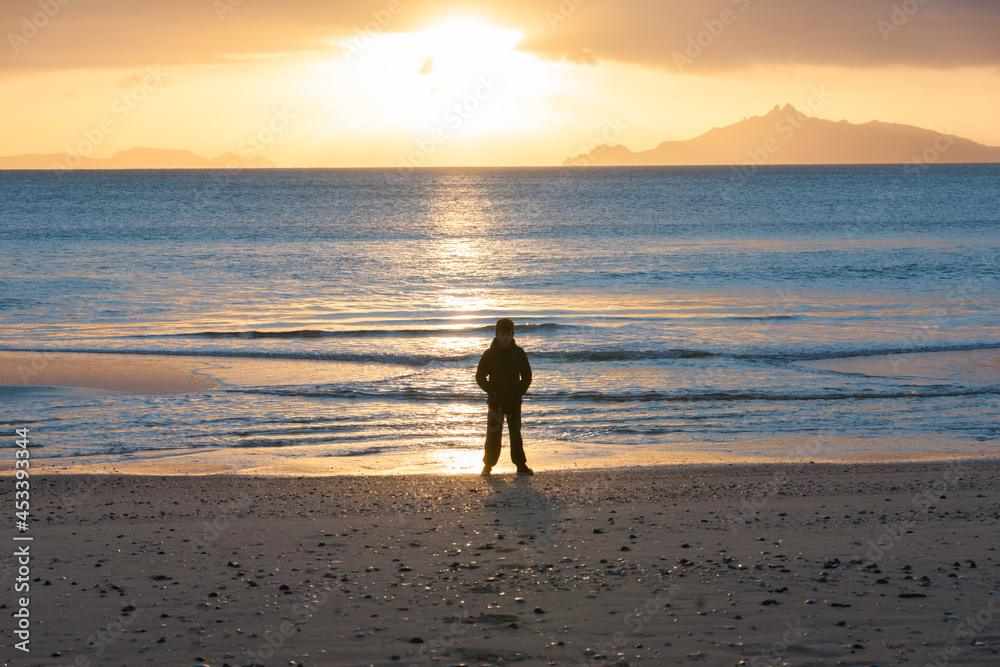 Naklejka premium Golden glow of sunrise across sea with silhouette of boy standing on beach on beach Waipu Cove
