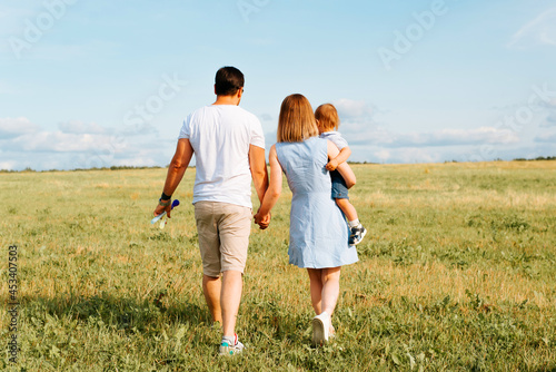 Family time together outdoors. Back view of young parents with child walking in field on sunny day at sunset, mom is carrying little boy in her arms, couple is holding hands
