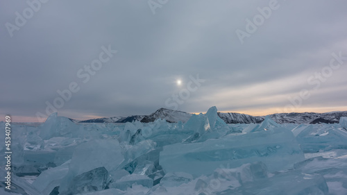 There is a block of turquoise hummocks on the surface of the frozen lake. Highlights on the edges. The setting sun in a cloudy sky. A mountain range in the distance. Baikal