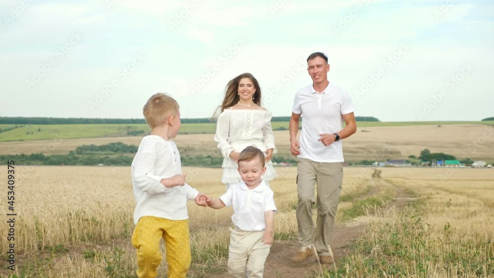 Parents and sons in white closes with joined hands run along wheat field ground road on hot summer day, against forest on horizon under blue sky Parents and sons in white closes with joined hands run along wheat field ground road on hot summer day, against forest on horizon under blue sky