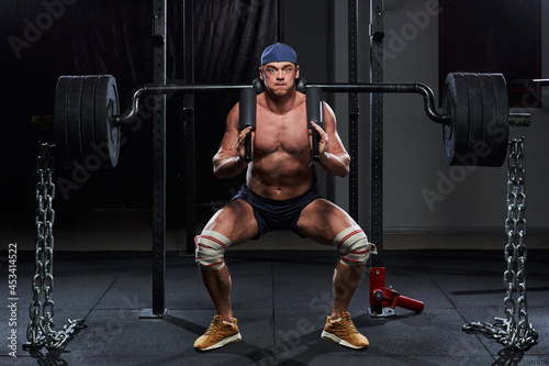 a young muscular man with a naked torso in the gym squats with a barbell on his shoulders, on which a lot of weights are hung. strength exercises in the gym. athlete