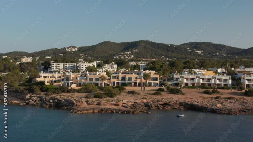 Aerial forward view of boats parked in a silent and calm sea with a young man standing at shore looking across with house and resort villa with greenery in Ibiza in Spain Aerial forward view of boats parked in a silent and calm sea with a young man standing at shore looking across with house and resort villa with greenery in Ibiza in Spain