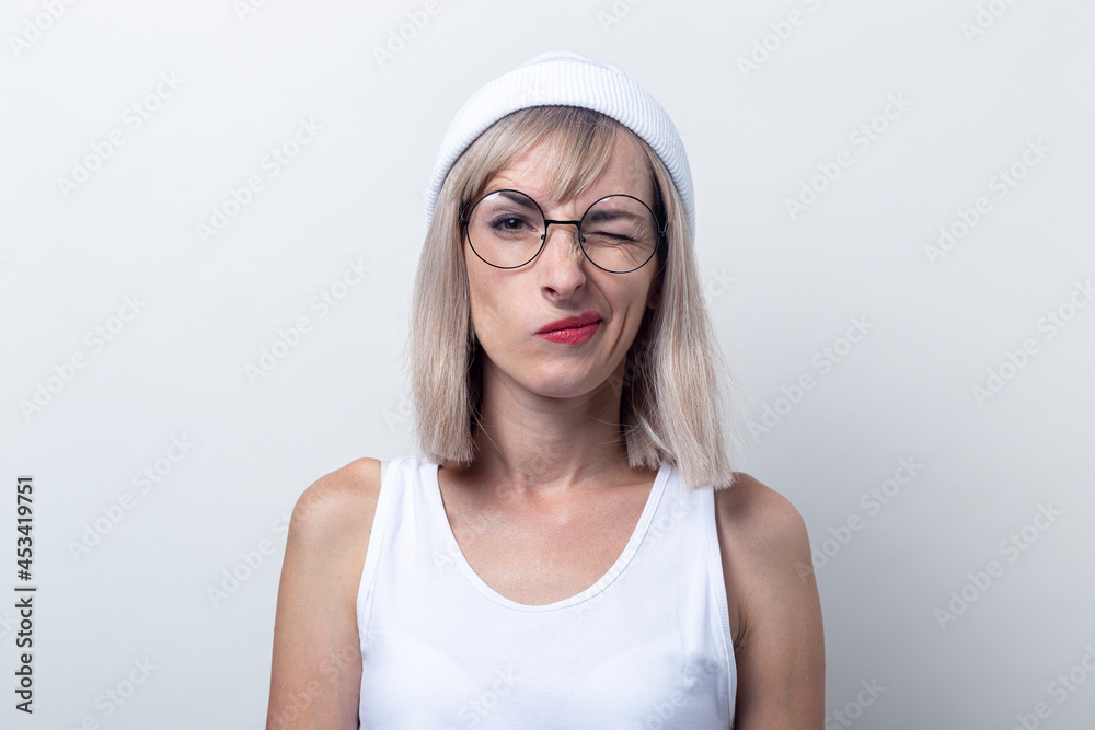 Blond young woman winking in glasses, a white hat on a light background Blond young woman winking in glasses, a white hat on a light background