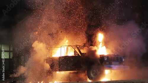 The car explodes and the hood and roof are torn off. A black SUV is torn apart by a huge orange flame of an explosion, the night on a city street. The car explodes from a bomb or a shell hit.