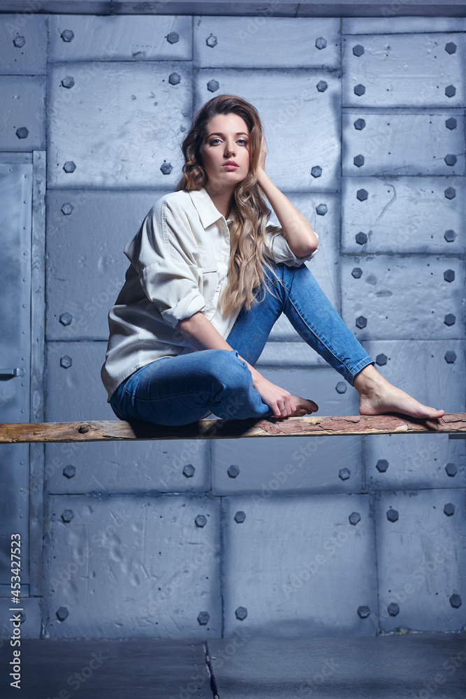 Portrait of young beautiful blonde woman is sitting on a board on metal wall background.