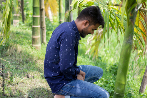 A young man is sitting To urinate with a small beard to wear a dark blue shirt and his behind green nature background blur.
