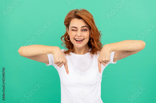 winking redhead girl with curly hair pointing finger down, copy space, sales and discounts.