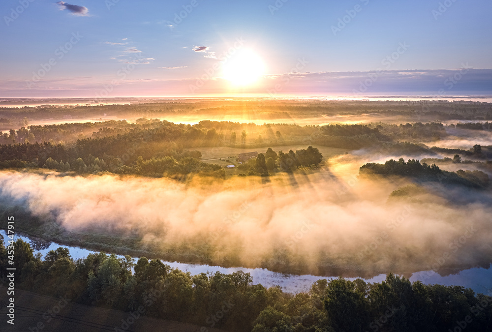 Light rays trough thick fog over a river in a misty summer morning. Rural landscape covered with pine forest and farm land.